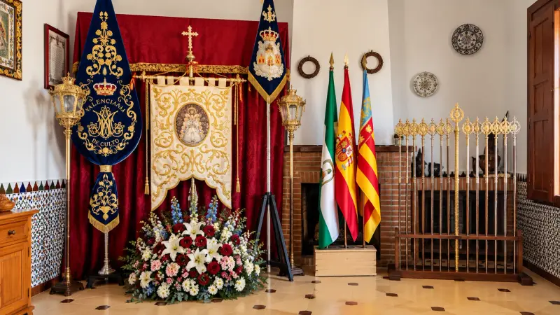 Simpecado y Estandarte de la Hermandad Valenciana de Culto, con sus varas, las banderas de Andalucía, España y la Comunitat Valenciana y un adorno floral, en la casa de hermandad